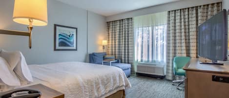 A hotel room with a bed, a desk with a television, a chair, a small table with a lamp, and a window with curtains.