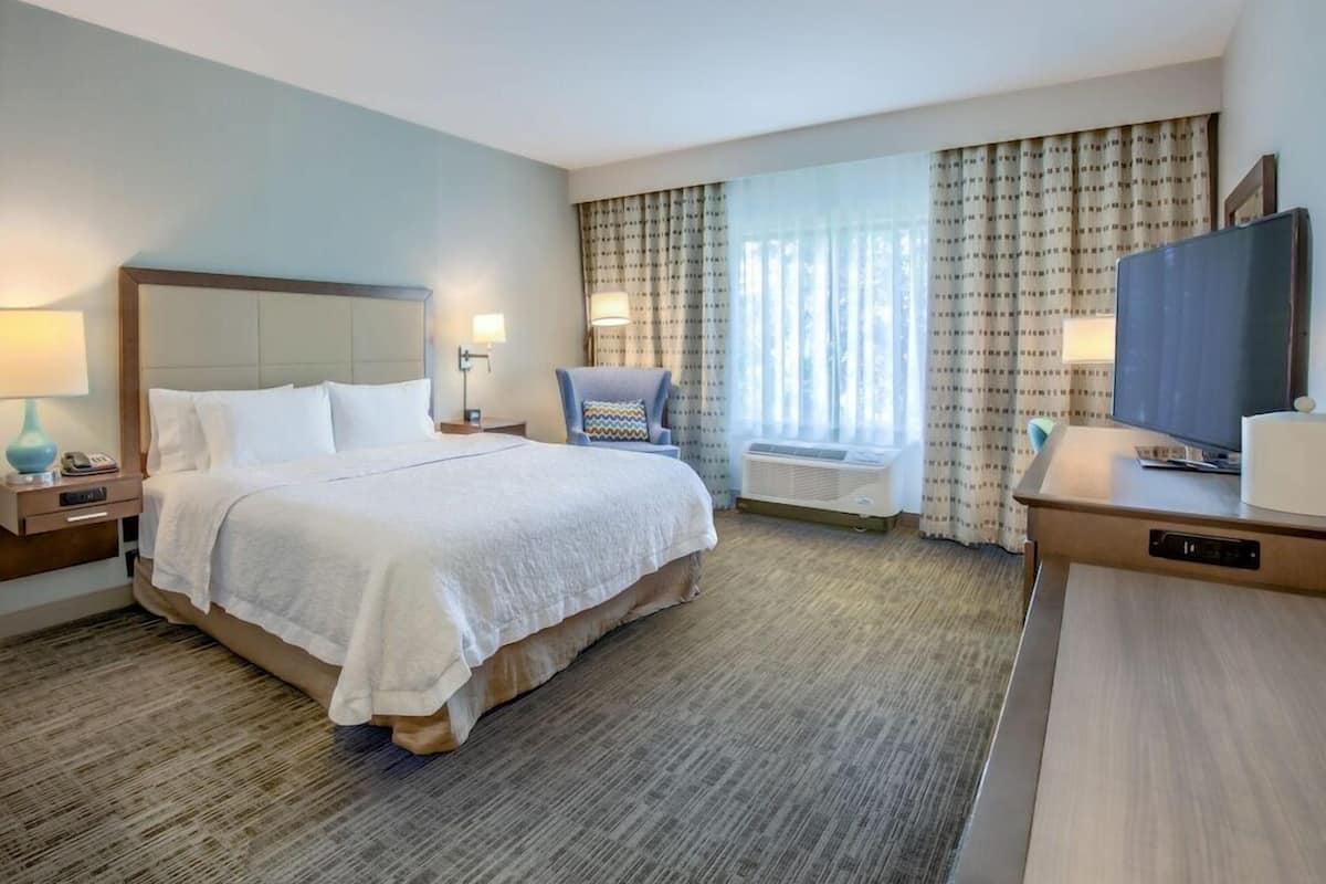 A hotel room with a bed, bedside tables, a chair, a desk, a television, and a window with curtains.