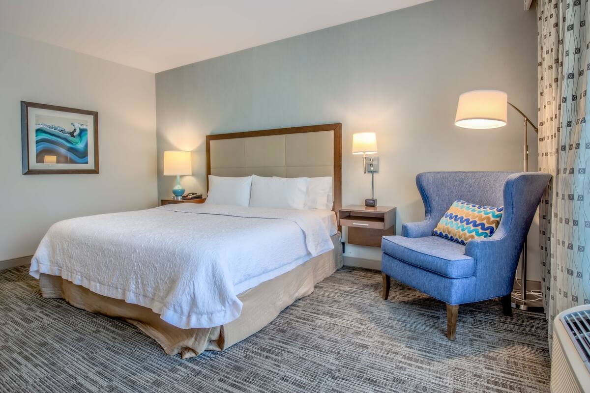 A hotel room with a large bed, a chair, a nightstand, a lamp, and a wall-mounted phone.