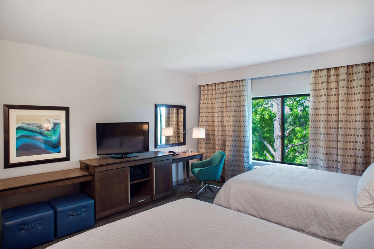 A hotel room with two beds, a desk with a television, a chair, and a large window with curtains.
