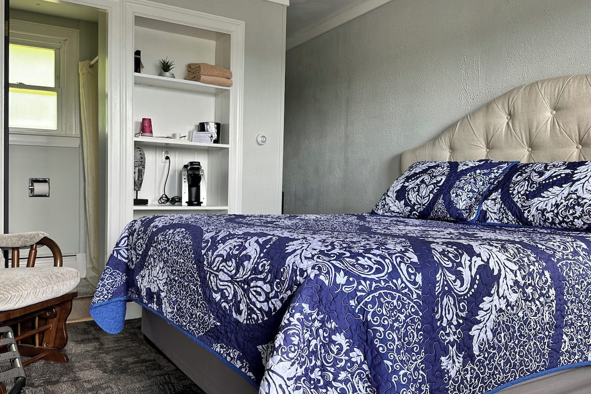 A bedroom with a bed, a chair, a shelf with various items, and a door leading to another room.
