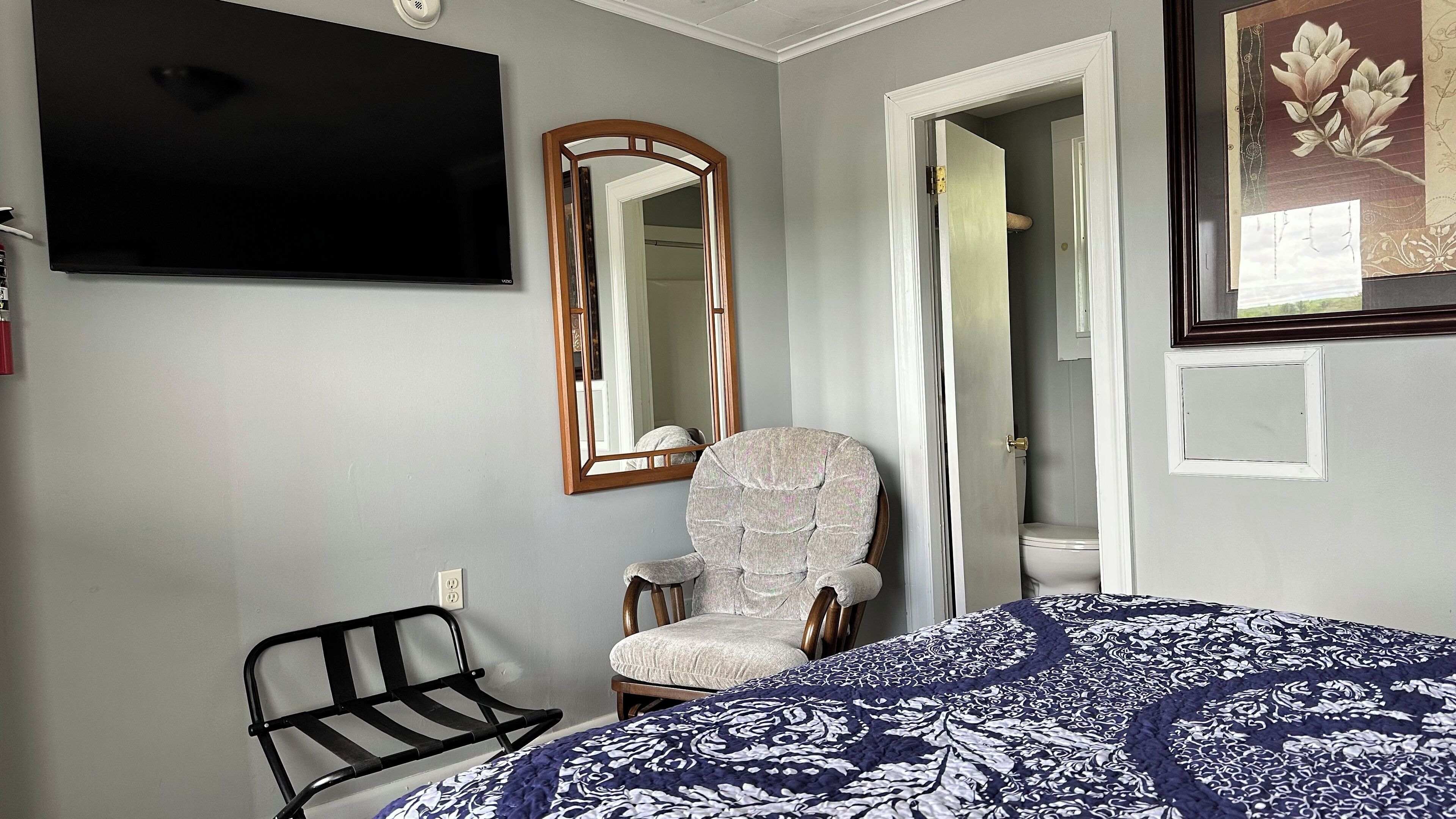 A bedroom with a bed, a chair, a television, a mirror, and a door leading to a bathroom.