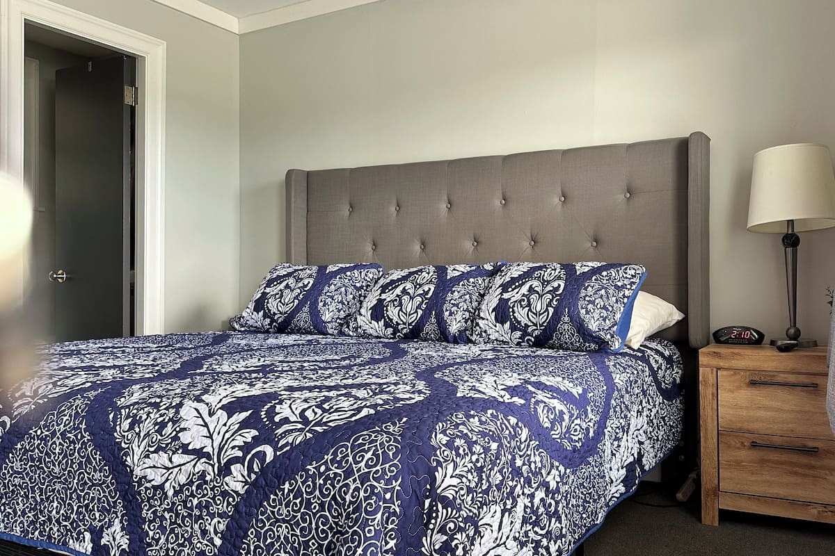 A bedroom with a tufted headboard, a patterned bedspread, a bedside table with a lamp, and a wooden nightstand.