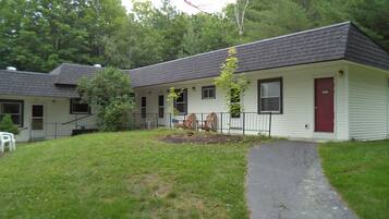 A single-story building with a dark roof and white siding, featuring a front porch with a bench and a small tree.