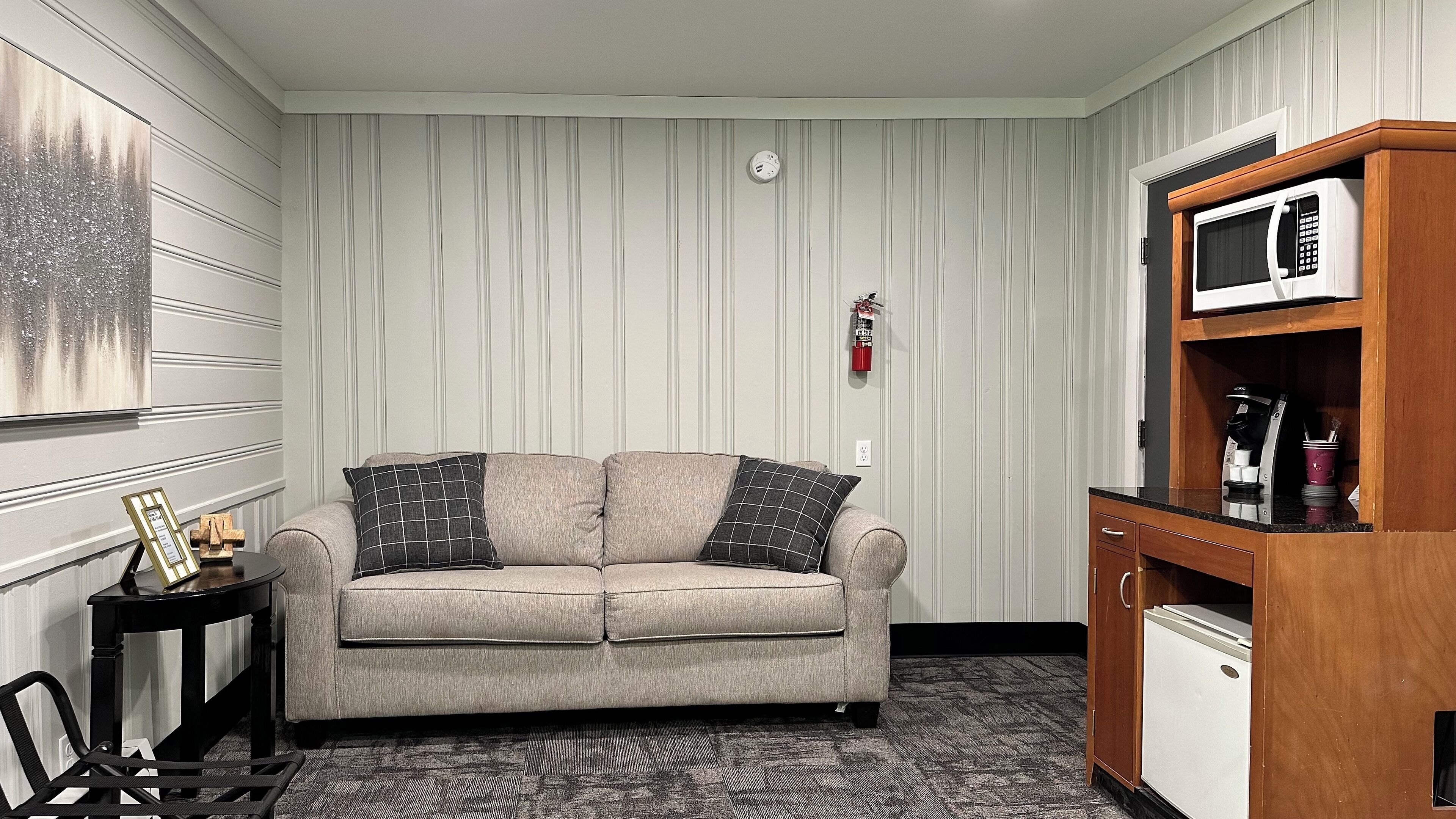 A living room with a beige sofa, two pillows, a small table with a picture frame, a coffee machine, and a microwave on a wooden cabinet.