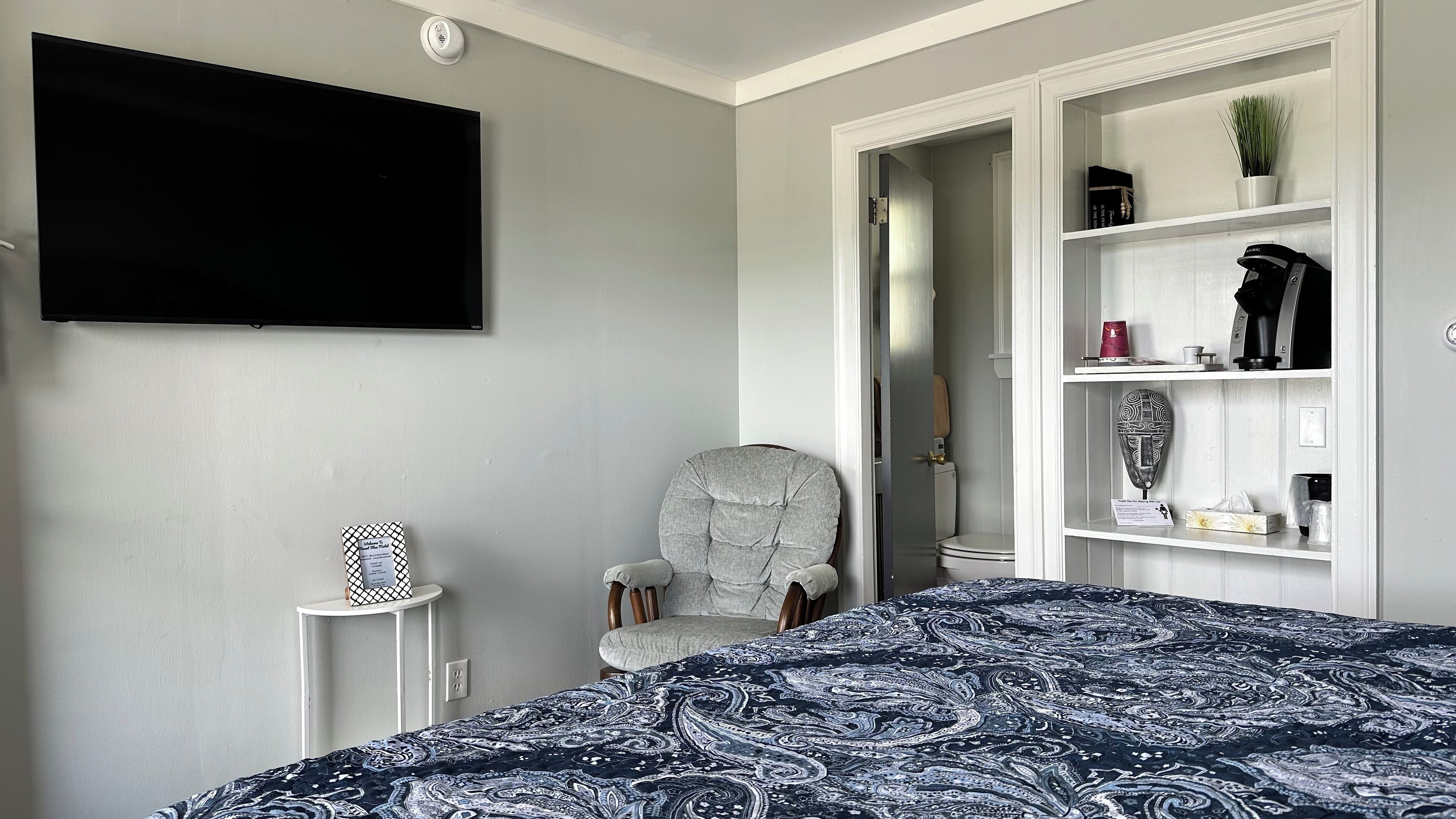 A bedroom with a bed, a chair, a TV, a shelf with various items, and a door leading to another room.