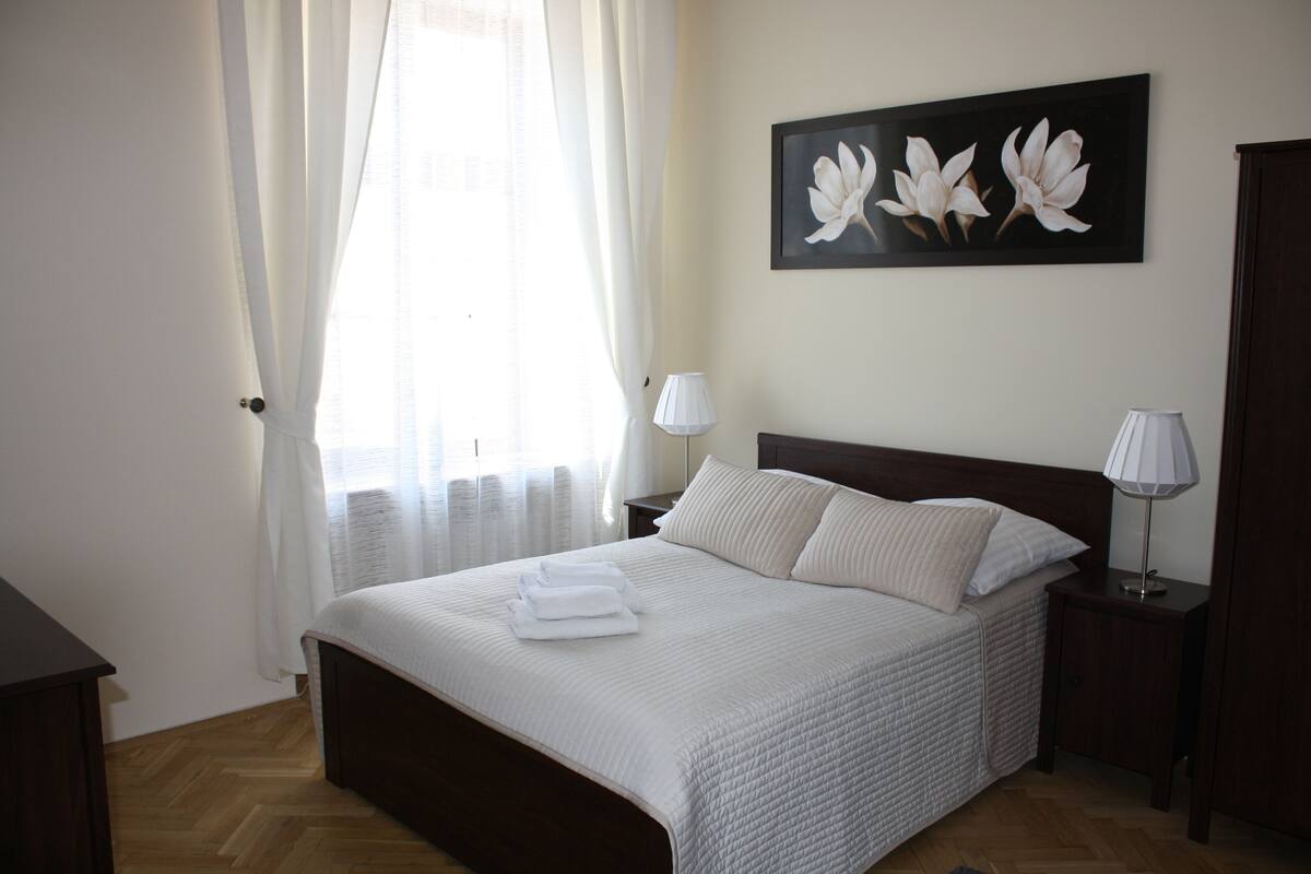 Ein ordentlich bezogenes Bett mit weißen Leinen und Kissen, ein Nachttisch mit einer Lampe und ein Blumengemälde an der Wand.