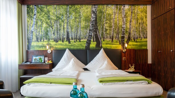 A hotel room with a large bed, green pillows, a nightstand with a lamp, and a wall mural of a forest. - Hotel Goldener Hirsch (Bayreuth)