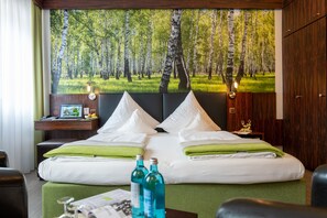 Habitación de hotel con una cama grande, almohadas verdes, una mesita de noche con lámpara y un mural en la pared de un bosque.