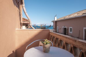 Ein Balkon mit einem weißen Tisch und einem Korbwicker-Korb voller Blumen, mit Blick auf eine Küstenlandschaft mit Gebäuden und blauem Himmel.