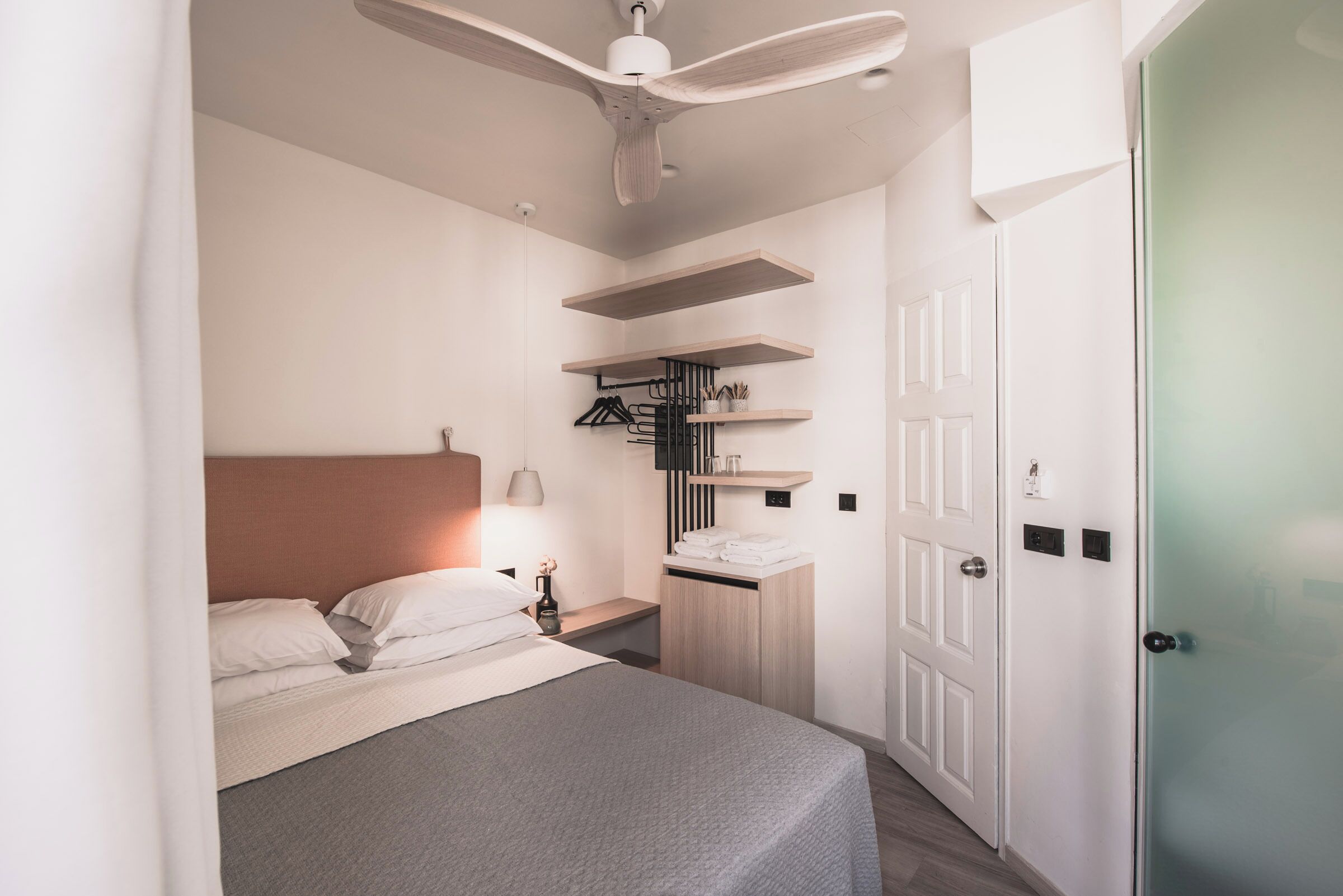 A modern bedroom with a bed, bedside table, and a ceiling fan. There are built-in shelves above the bed and a door to the right.