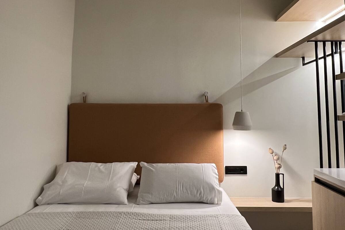 A modern bedroom with a large bed, a ceiling fan, a bedside table with a lamp, and a view of the outside.