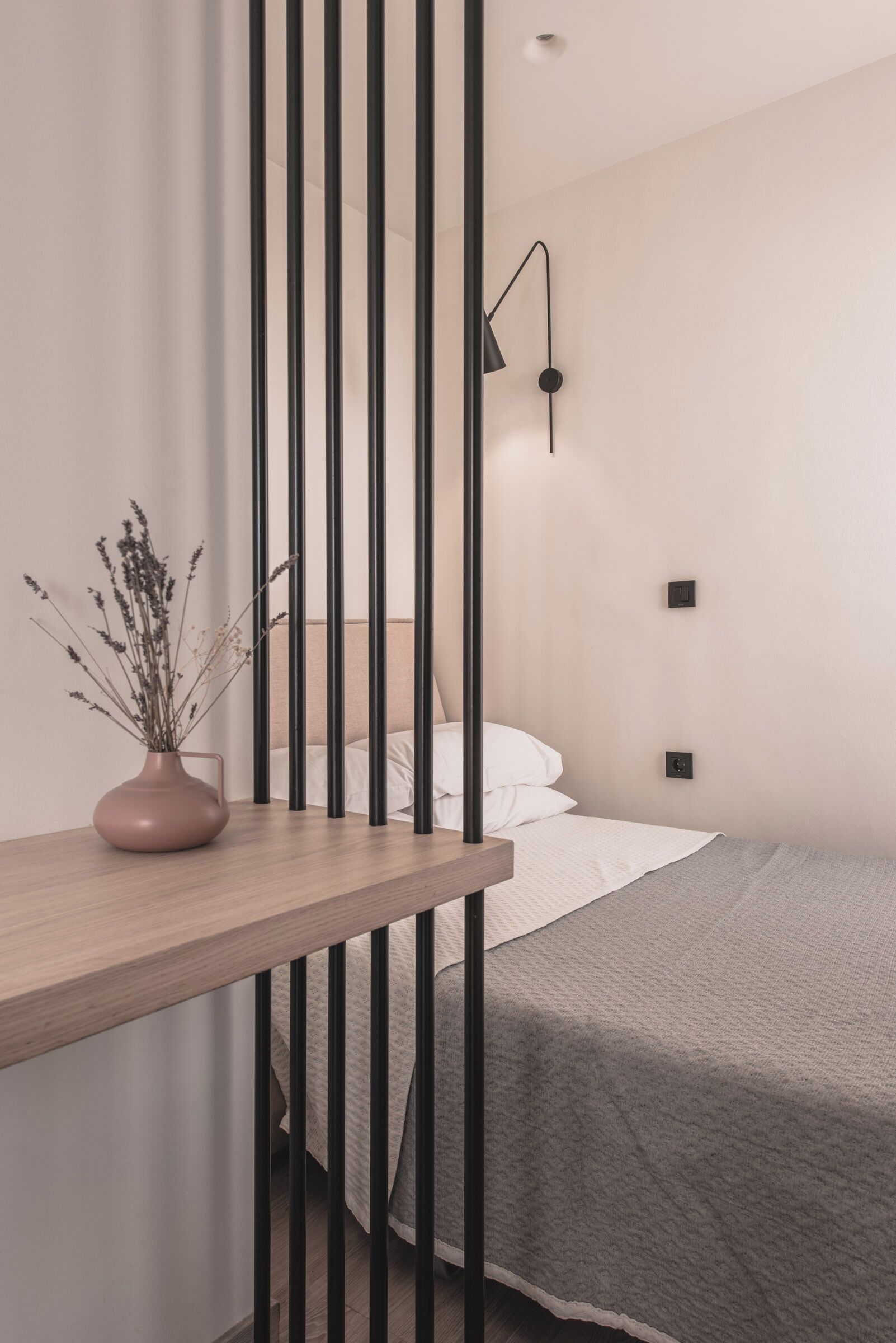 A modern bedroom with a wooden bedside table, a vase with dried flowers, a bed with a grey blanket, and a black wall-mounted lamp.