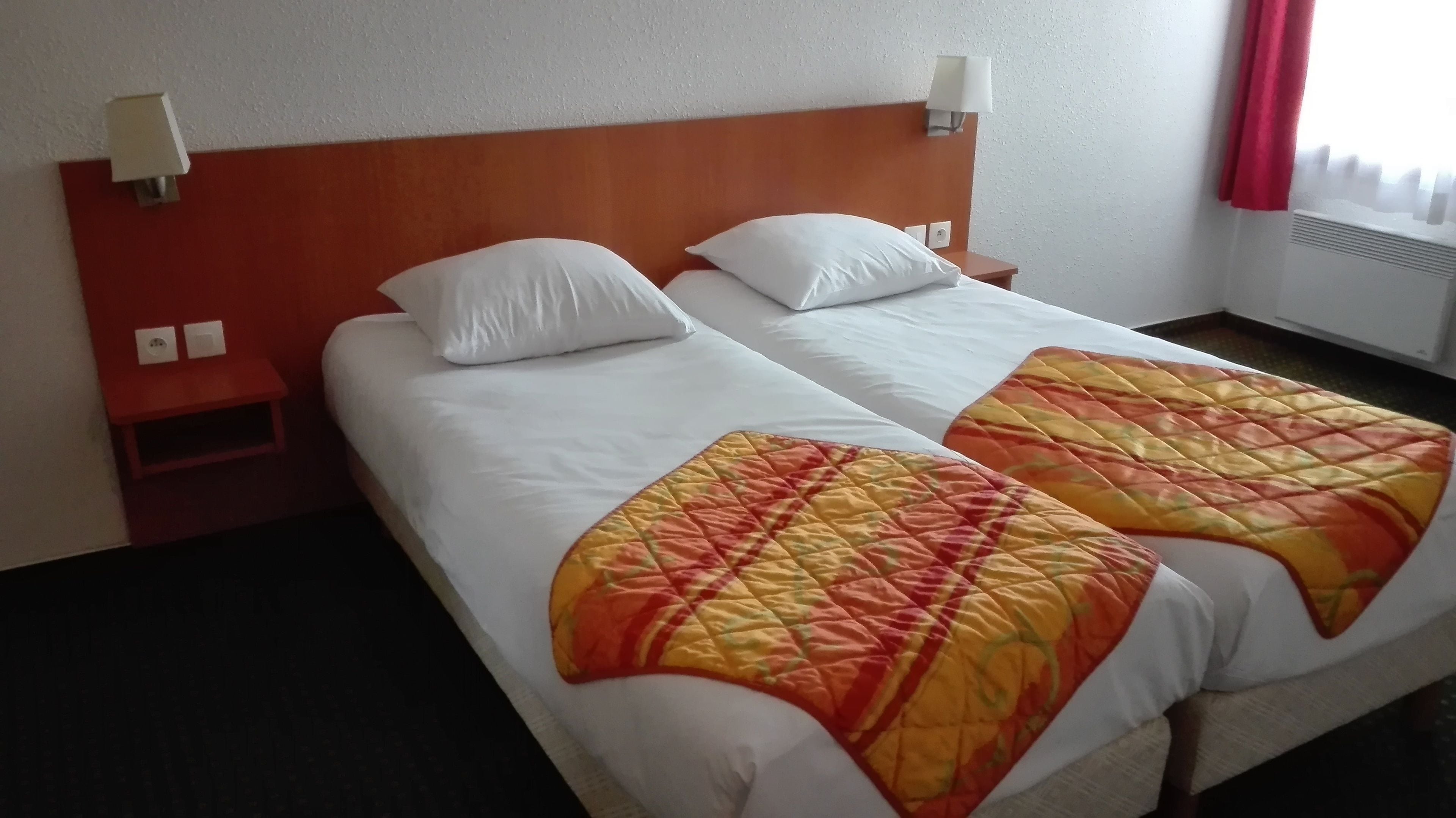Uma cama de casal com roupa de cama branca e duas almofadas, uma cabeceira de madeira, uma mesa de cabeceira com abajur e uma janela com cortinas.