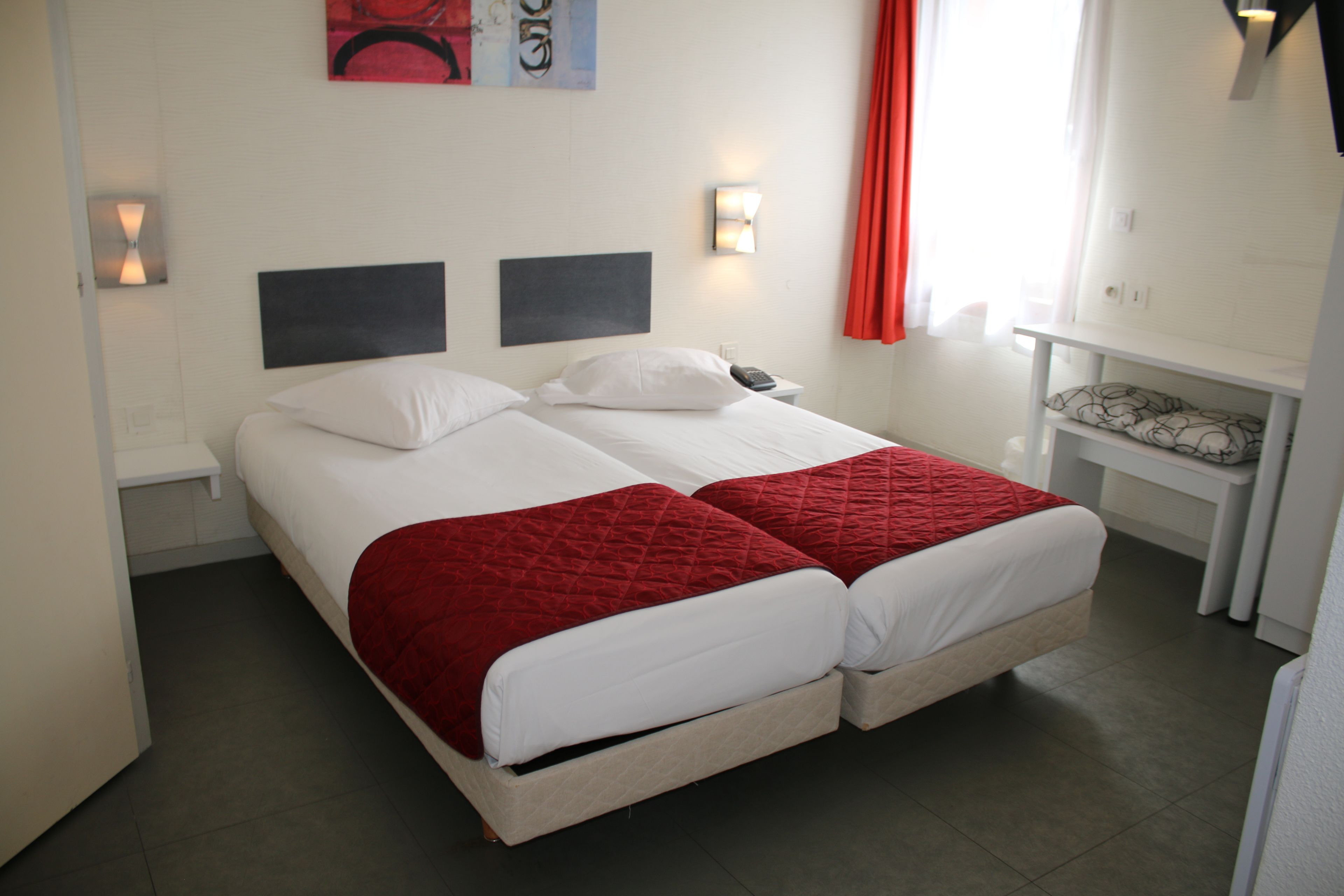 Ein Doppelbett mit roter Steppdecke, weißen Laken und einem Kopfteil mit zwei schwarzen Paneelen. Ein Nachttisch mit Lampe und Telefon ist vorhanden.
