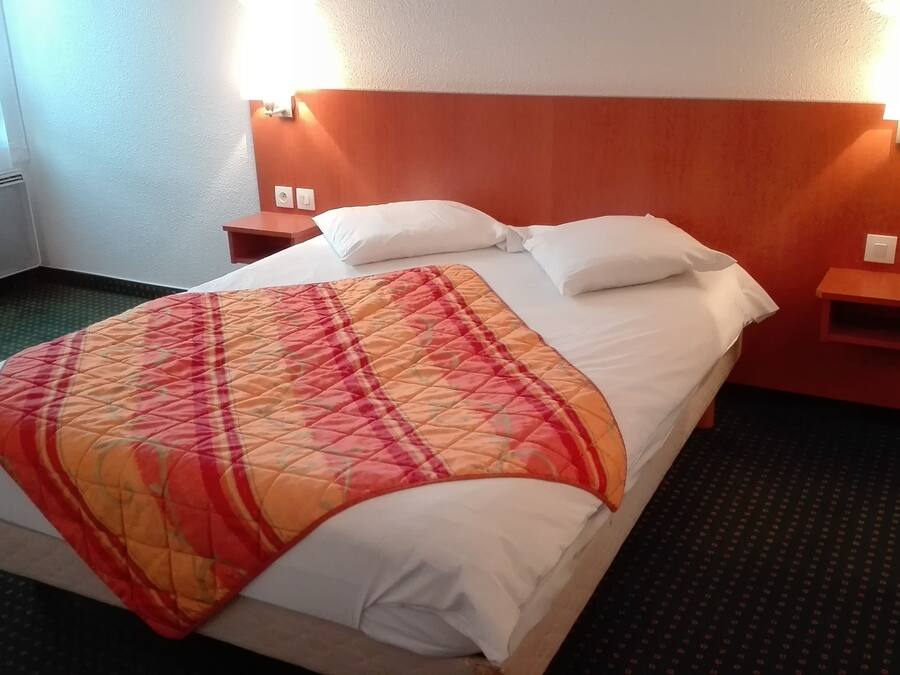 A hotel room with a bed, a bedside table, a nightstand, and a wall-mounted light fixture.