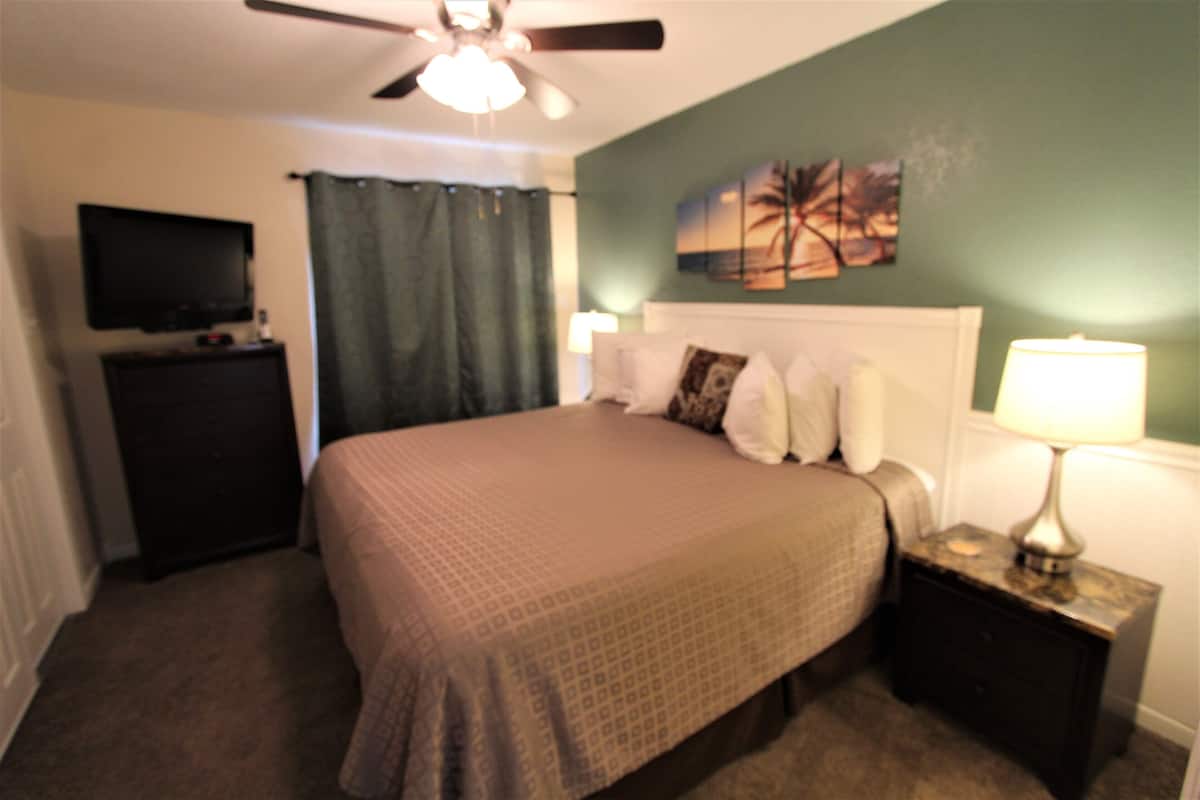 A bedroom with a bed, a television on a dresser, a ceiling fan, and a nightstand with a lamp.