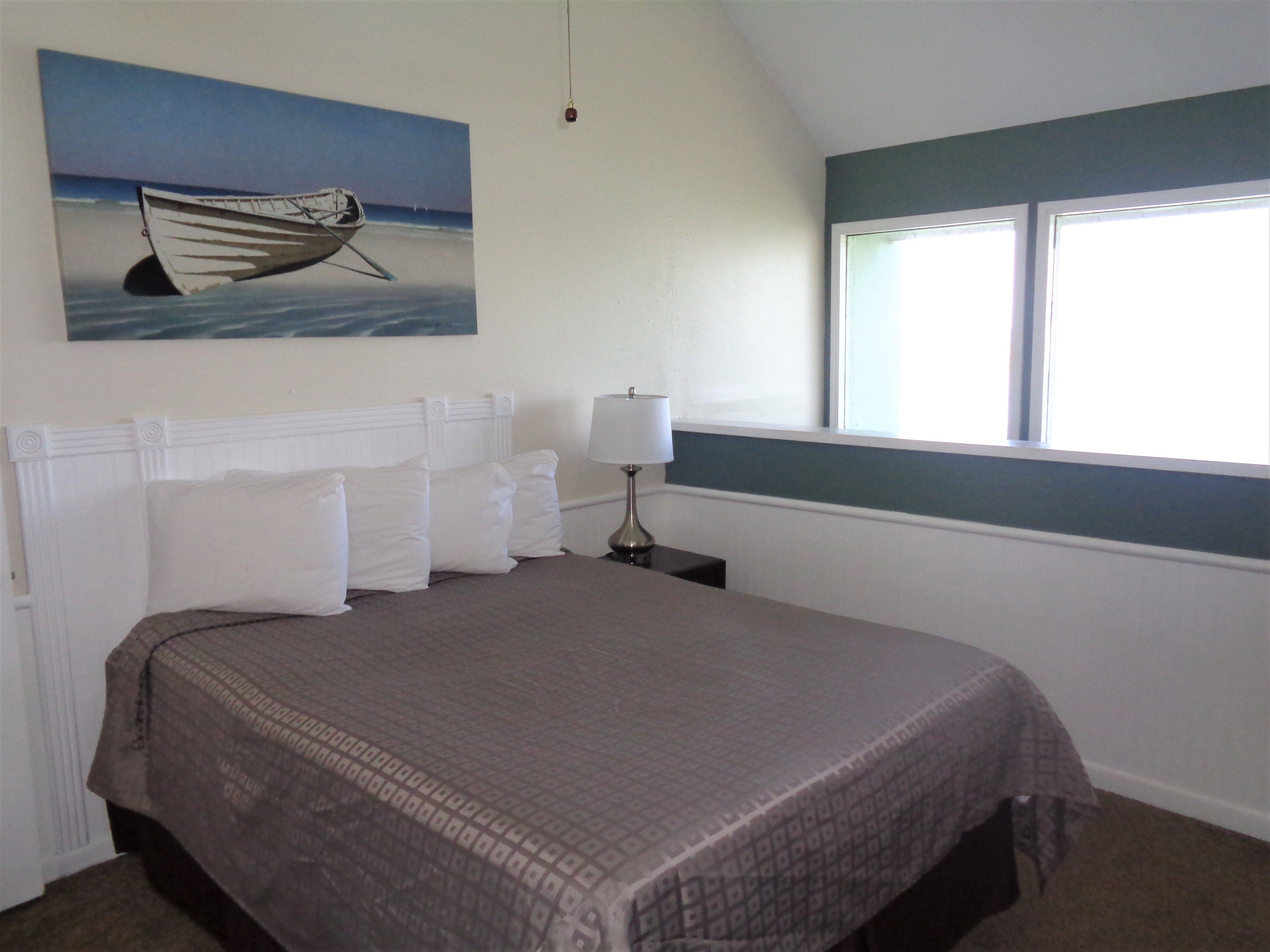 Un dormitorio con cama, cabecera blanca, un cuadro de un barco en la pared, una mesita de noche con lámpara y una ventana con persianas.