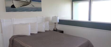 Un dormitorio con cama, cabecera blanca, un cuadro de un barco en la pared, una mesita de noche con lámpara y una ventana con persianas.