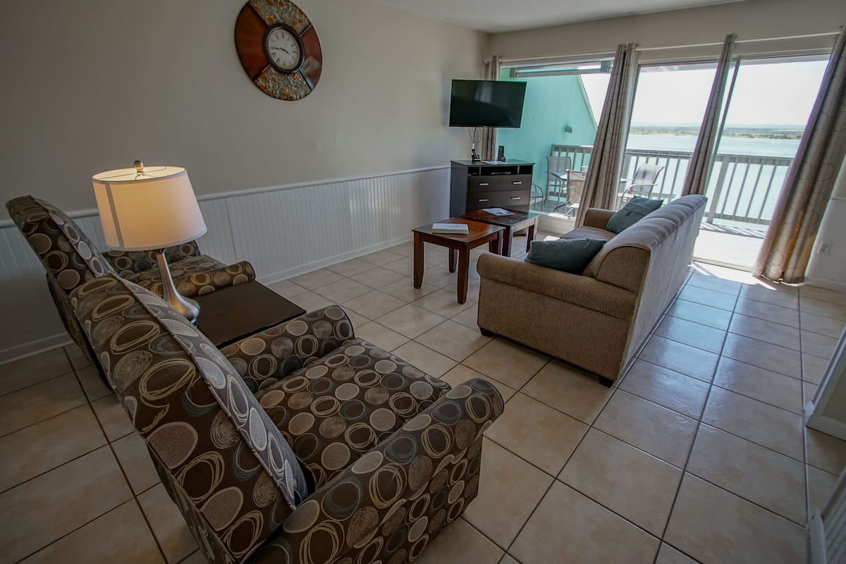 A living room with a sofa, a patterned armchair, a coffee table, a TV, and a balcony with a view.
