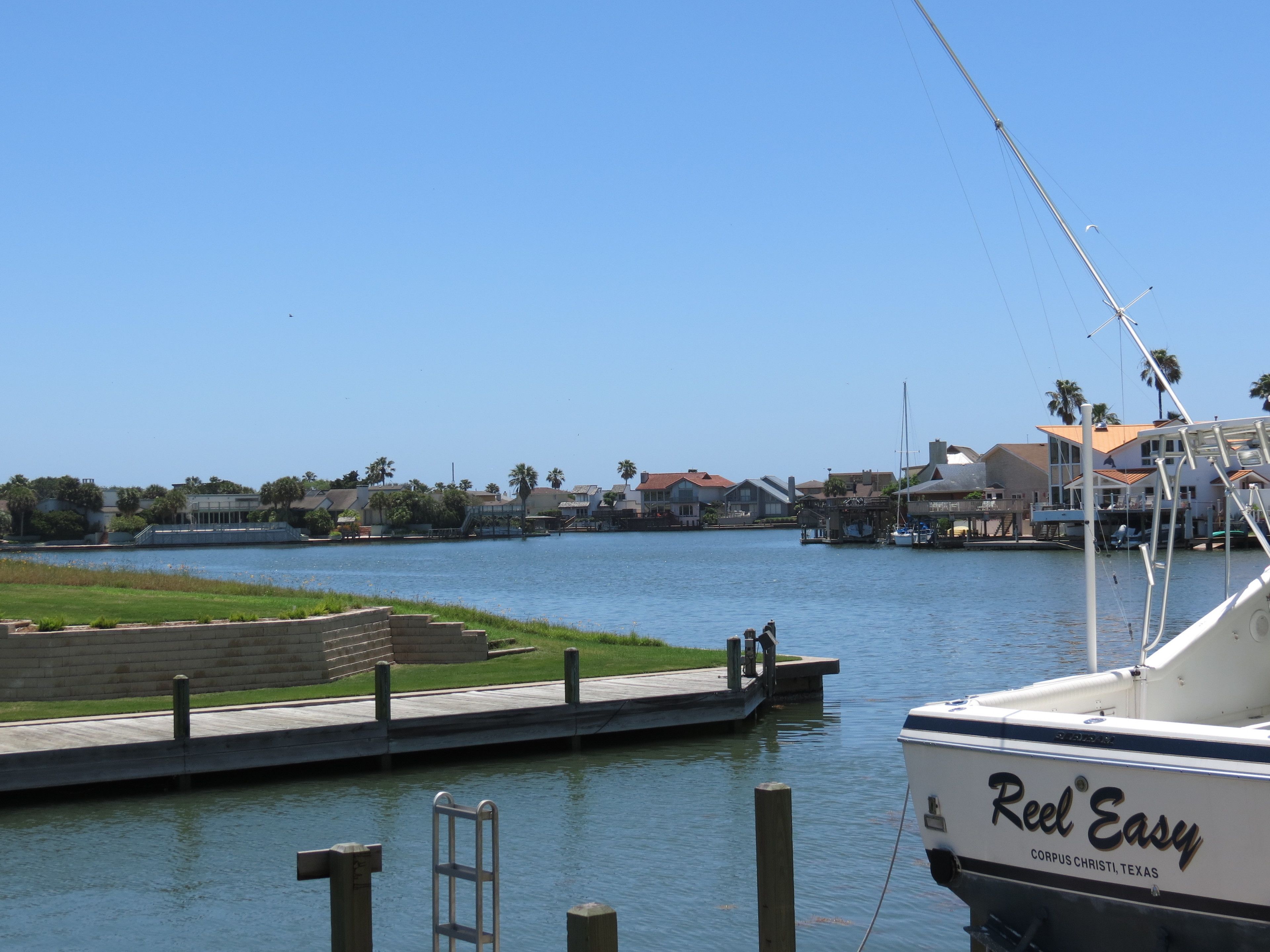 A boat docked at a marina with a clear view of the water and a distant shoreline with houses and palm trees.