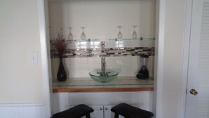 Une cuisine avec un évier en verre, deux tabourets, un mixeur et un vase contenant des plantes séchées.