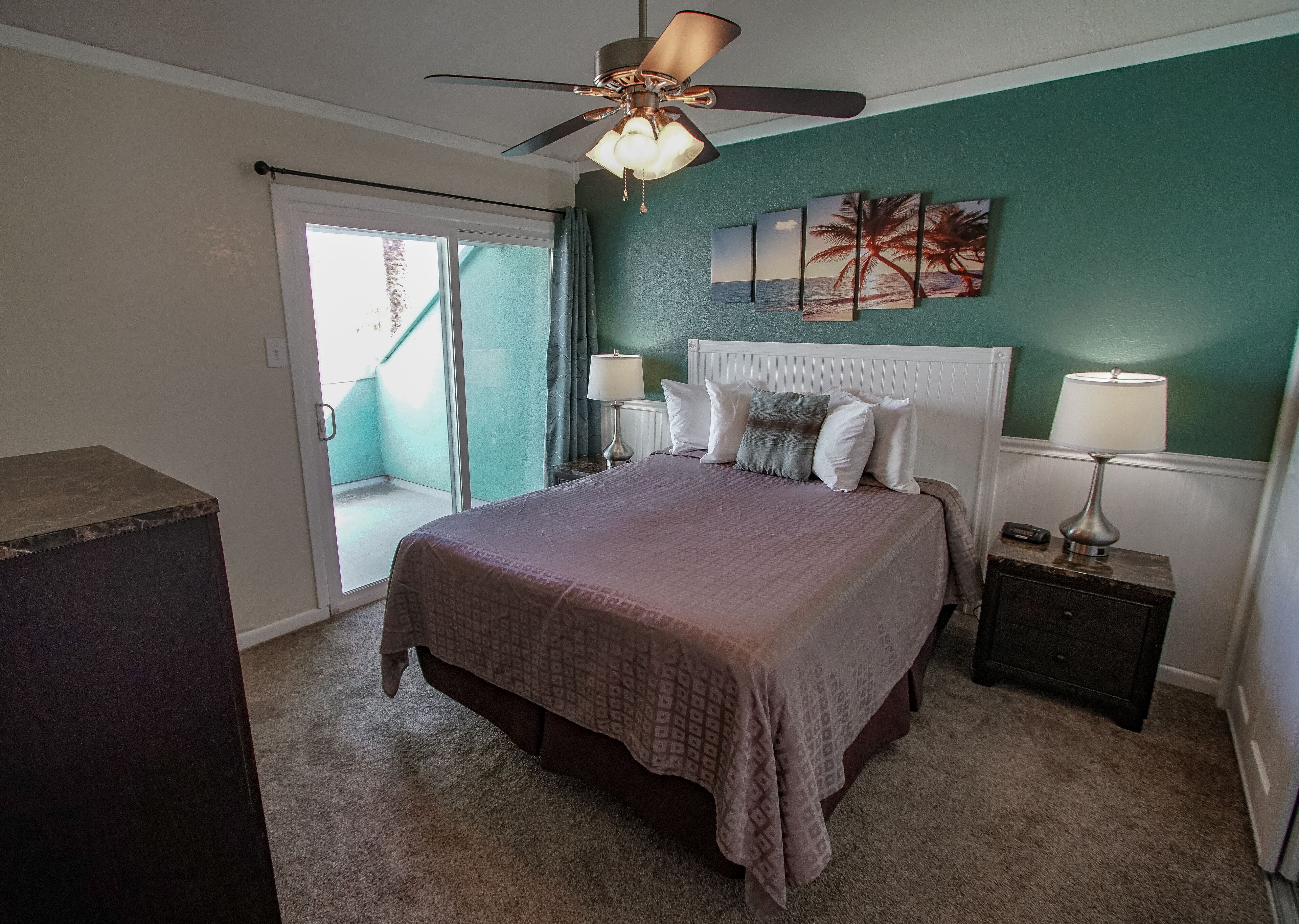 A bedroom with a bed, a ceiling fan, a nightstand, a lamp, and a view of the outside.