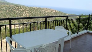 A balcony with a white table and two white chairs, overlooking a hilly landscape with a view of the sea. - Socrates Apartments (Neo Chorio)