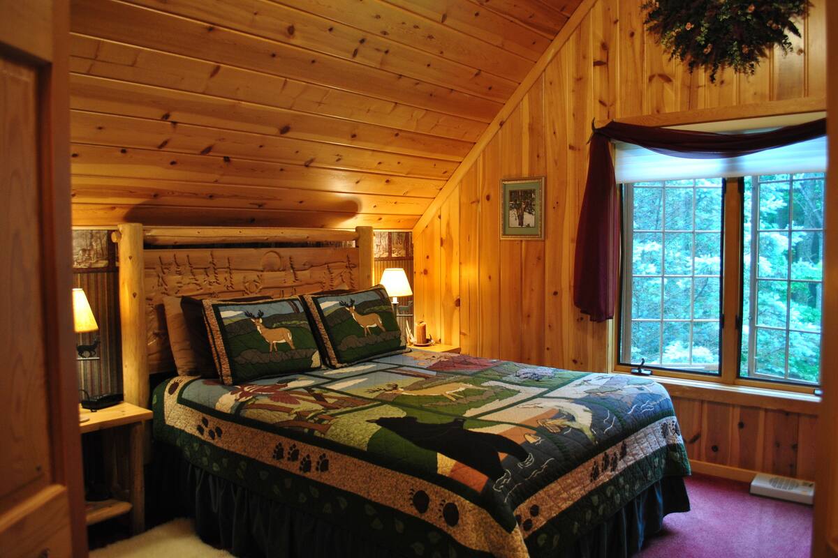 A log cabin bedroom with a large bed, wooden walls, a window with a view of trees, and a quilt with animal prints.