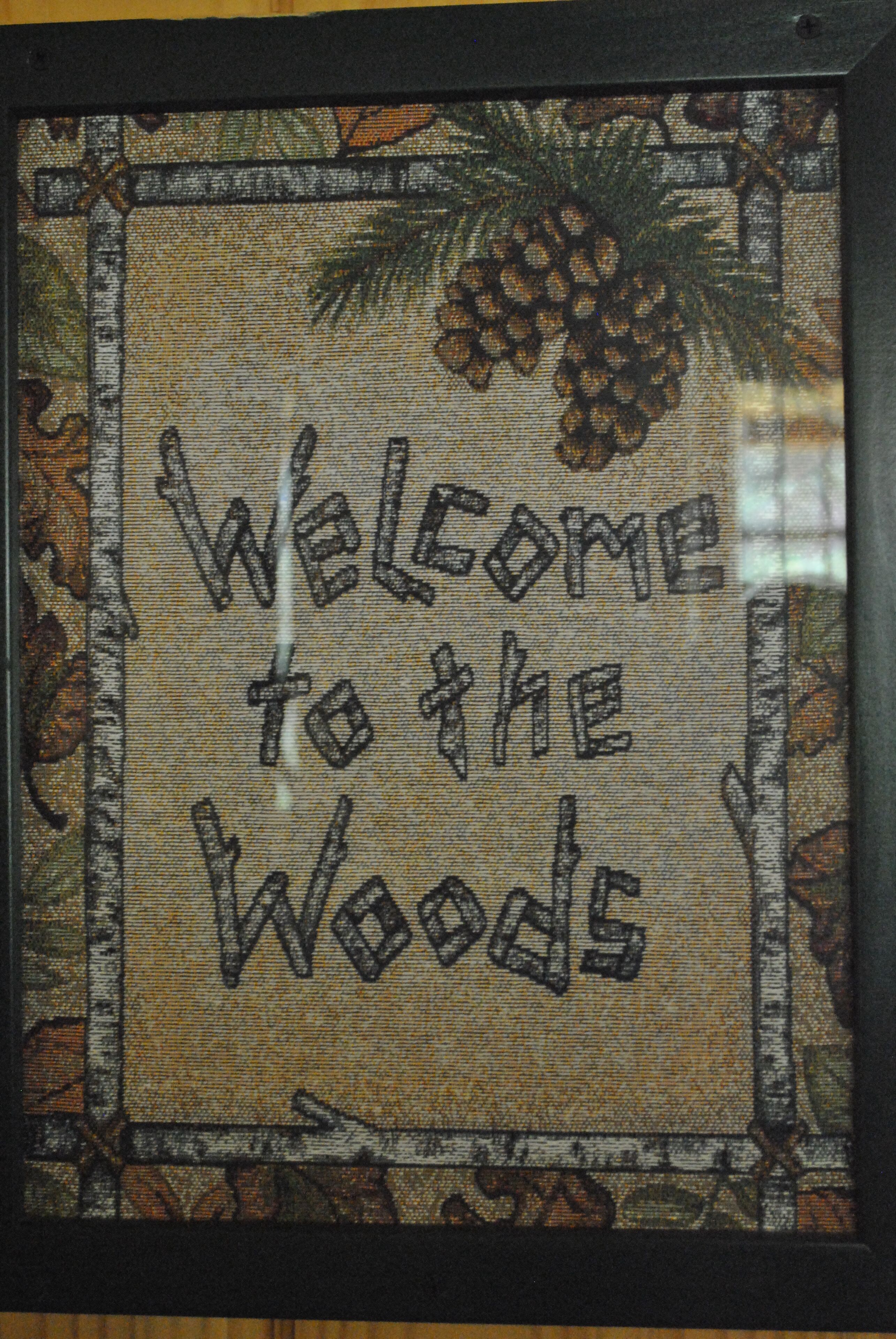 Placa de boas-vindas emoldurada com o texto "Welcome to the Woods" e elementos decorativos como pinhas e folhas.