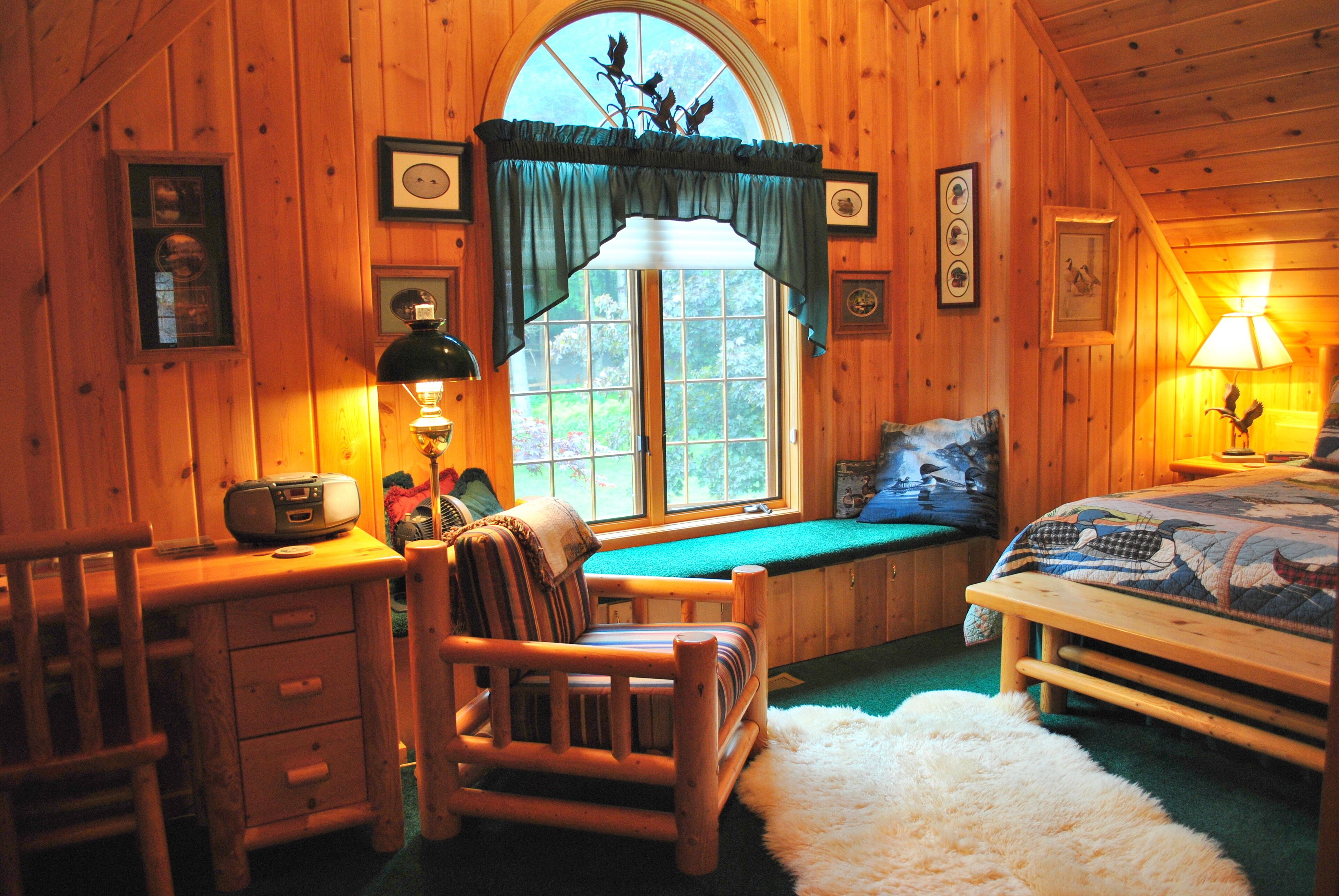 A cozy cabin room with a wooden interior, a large window with curtains, a bed with a quilt, a desk with a lamp, and a chair.