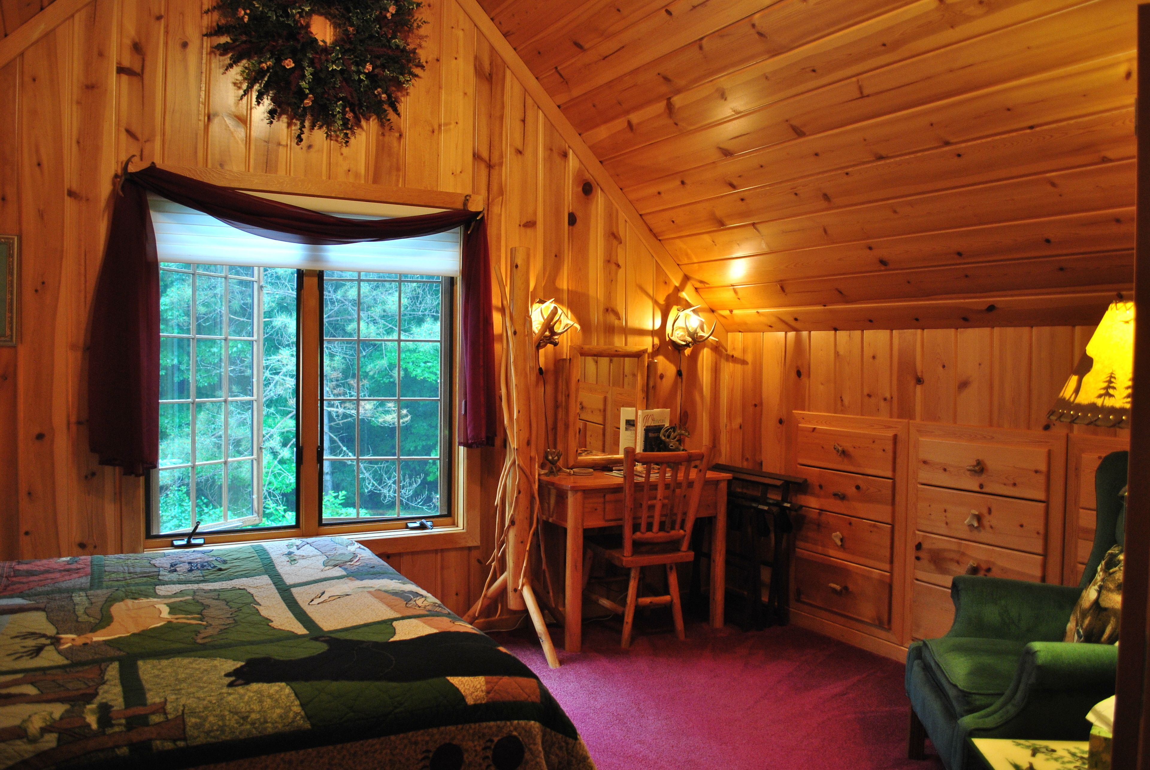 A wooden cabin room with a bed, a desk, a chair, and a dresser.