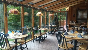 A cozy indoor dining area with wooden tables and chairs, large windows, and a Christmas tree. - Horton Creek Inn B&B (Charlevoix)