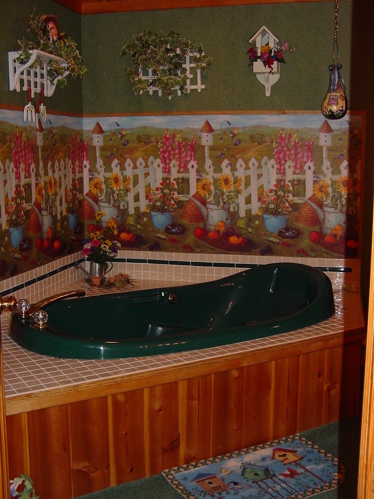 A bathroom with a green bathtub, a white picket fence mural, and a hanging flower vase.