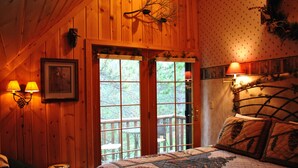 A wooden cabin bedroom with a large bed, two bedside lamps, a framed picture, and a view through a door to an outdoor deck. - Horton Creek Inn B&B (Charlevoix)