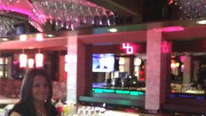Zona de bar con una mujer sentada en la barra, una pantalla de televisión y varios objetos de bar.