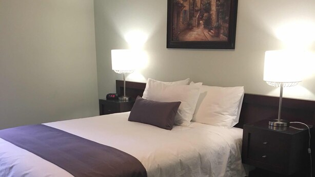 Una cama bien hecha con ropa de cama blanca y oscura, dos lámparas de noche, un cuadro enmarcado en la pared y un sillón de cuero.