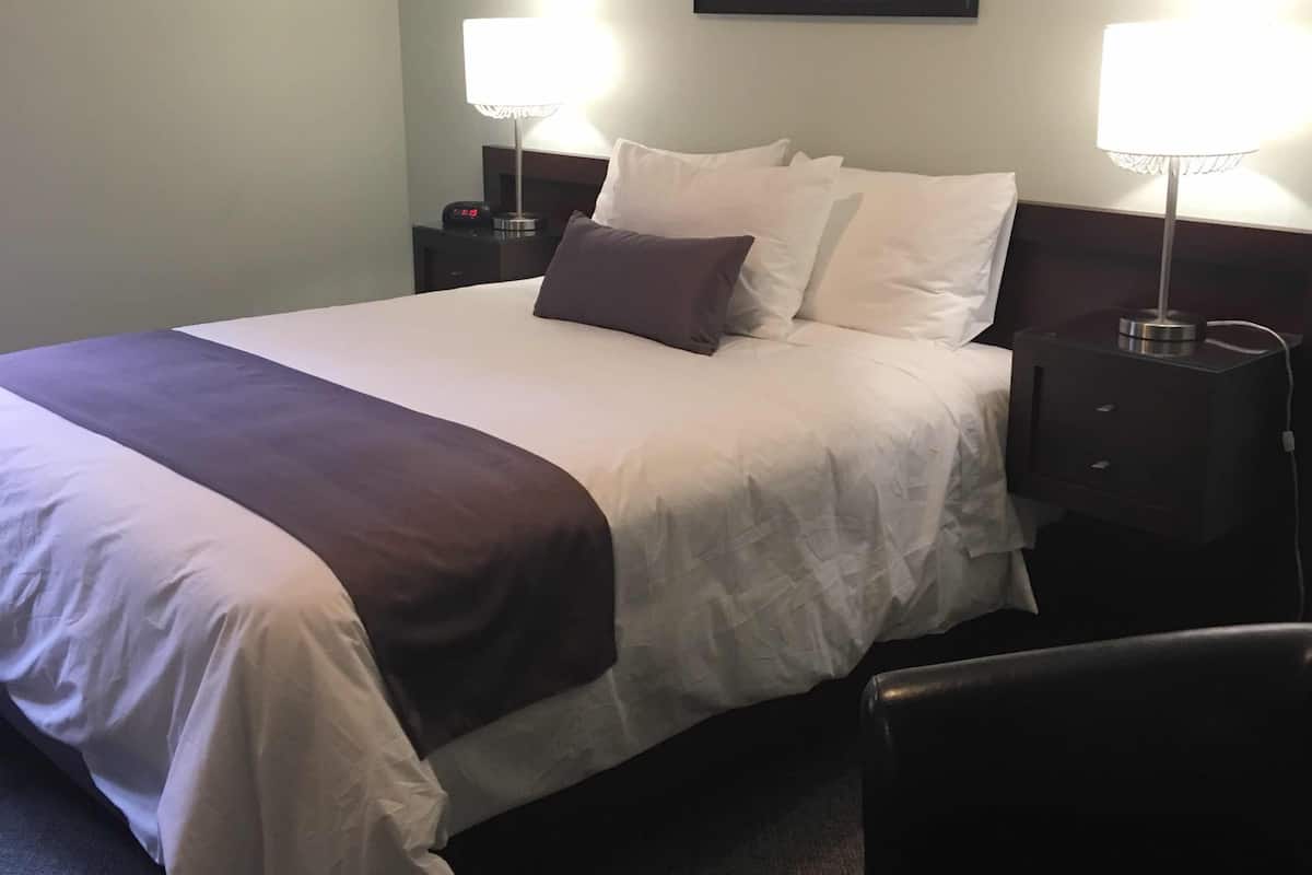 A neatly made bed with white and dark-colored linens, two bedside lamps, a framed painting on the wall, and a leather chair.