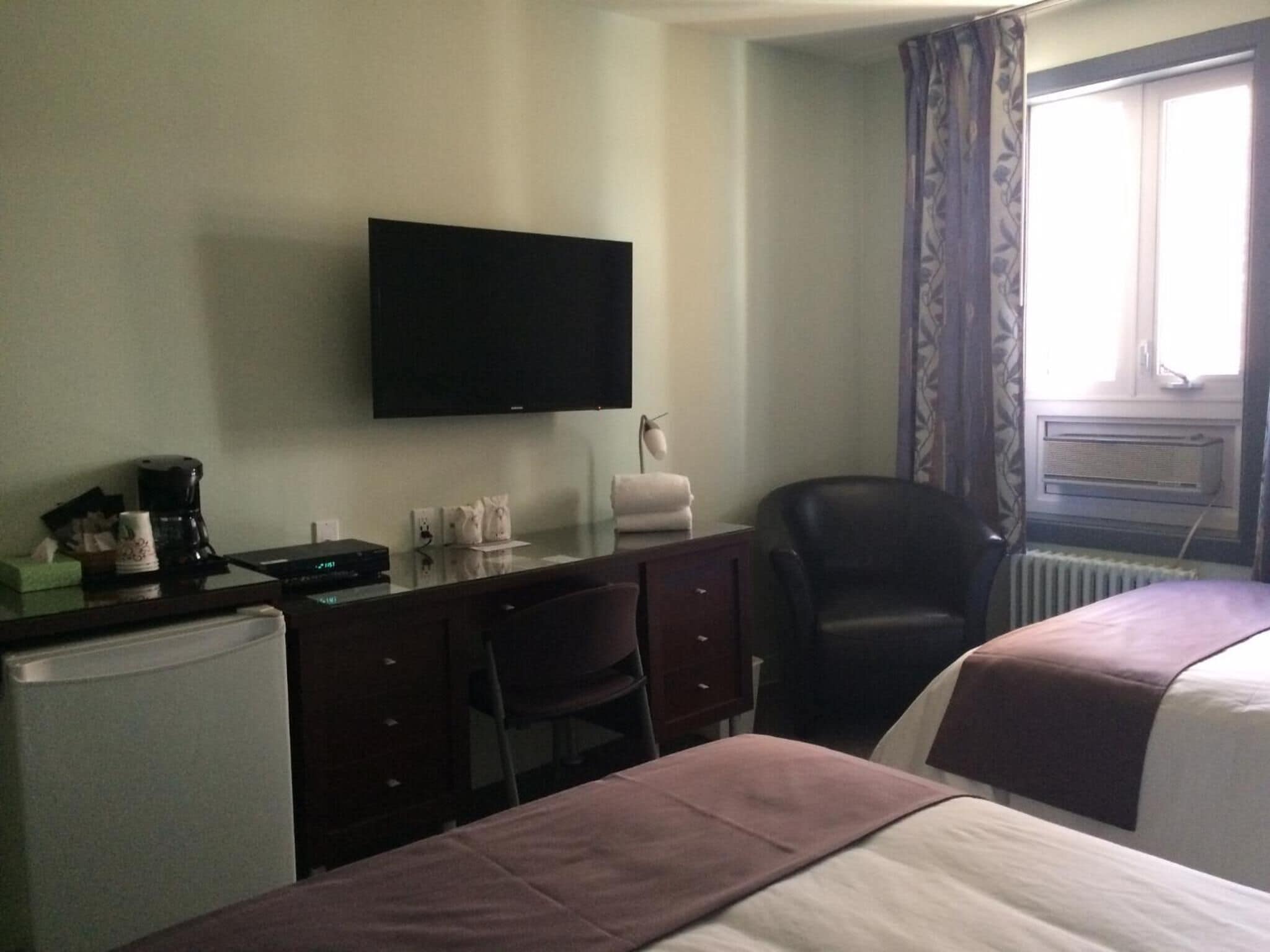 Quarto de hotel com cama, escrivaninha com computador, cadeira, televisão e janela com cortinas.