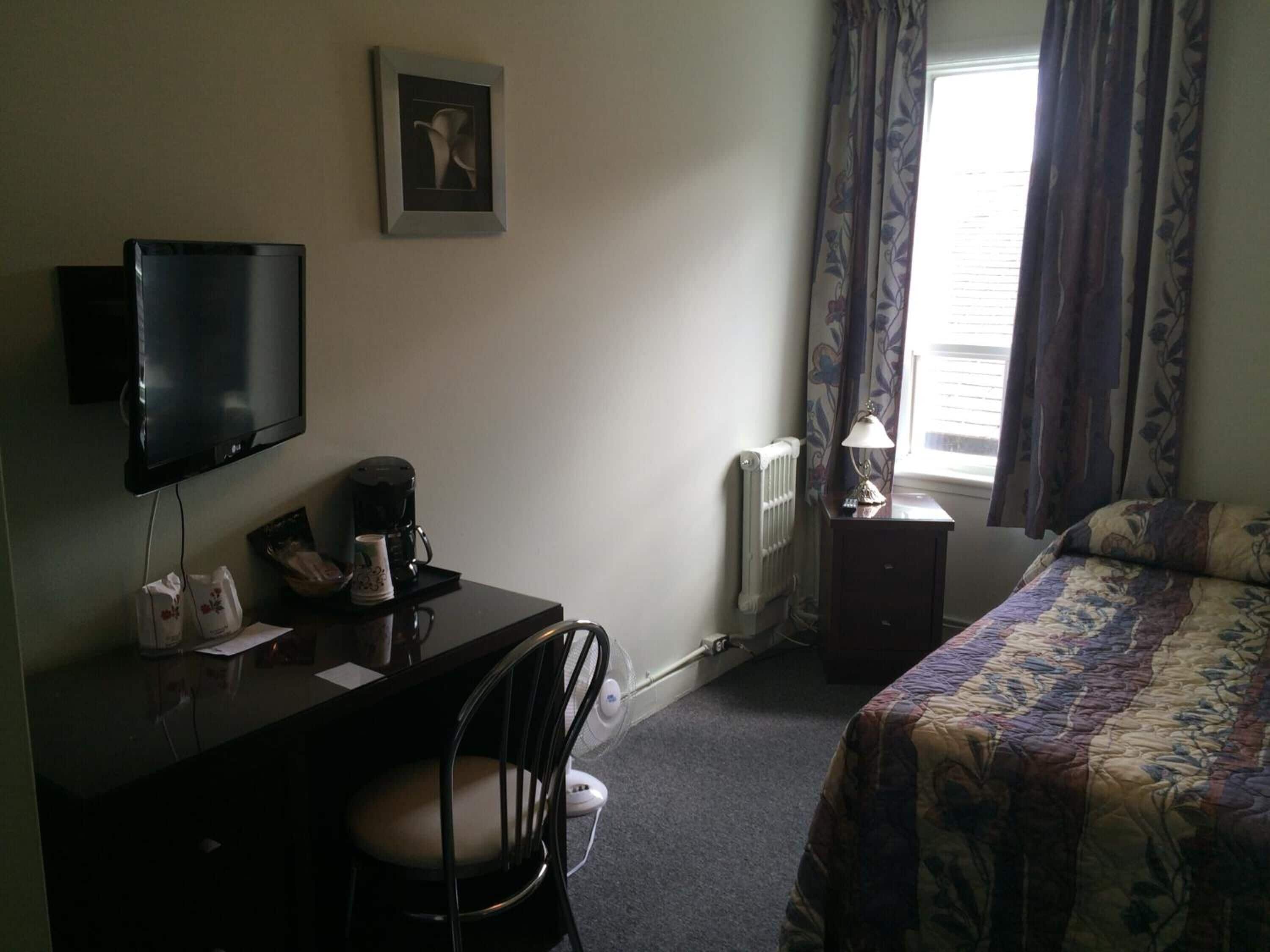 A hotel room with a bed, a desk with a chair, a television, a radiator, and a window with curtains.