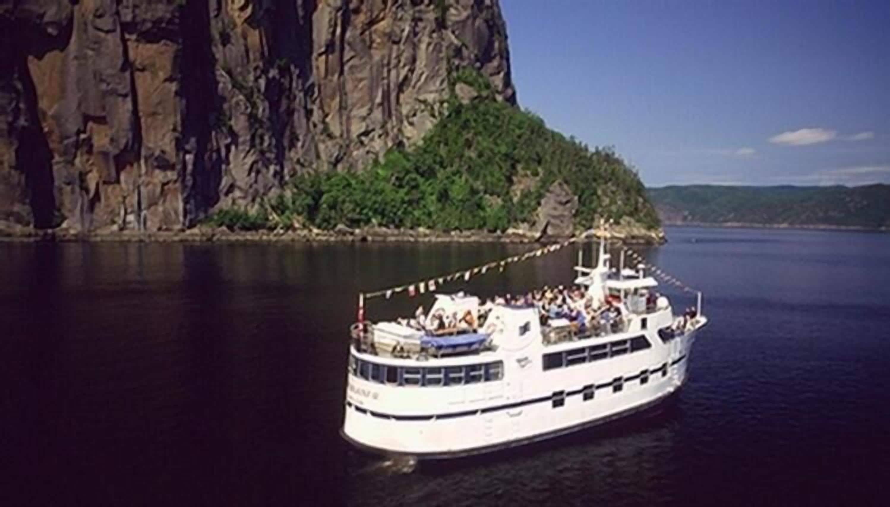 A large white boat with multiple decks, docked near a rocky shoreline with greenery.