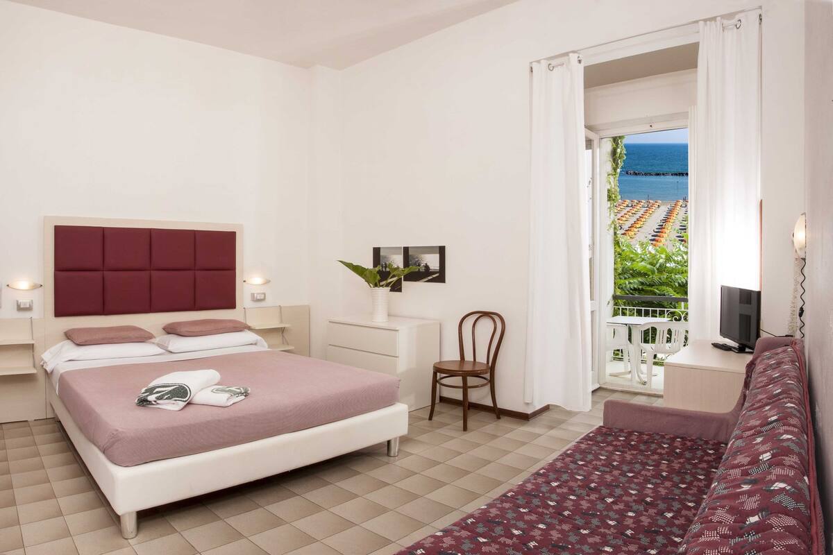 Une chambre avec un lit, des tables de chevet, une chaise, un bureau, une télévision et un balcon avec vue sur la mer.