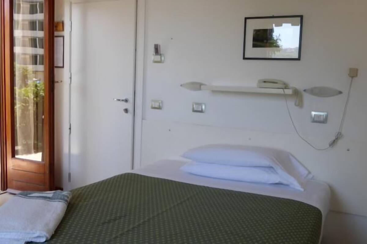 Une chambre avec un lit, une petite table de chevet, un téléphone fixé au mur et une fenêtre avec des stores.