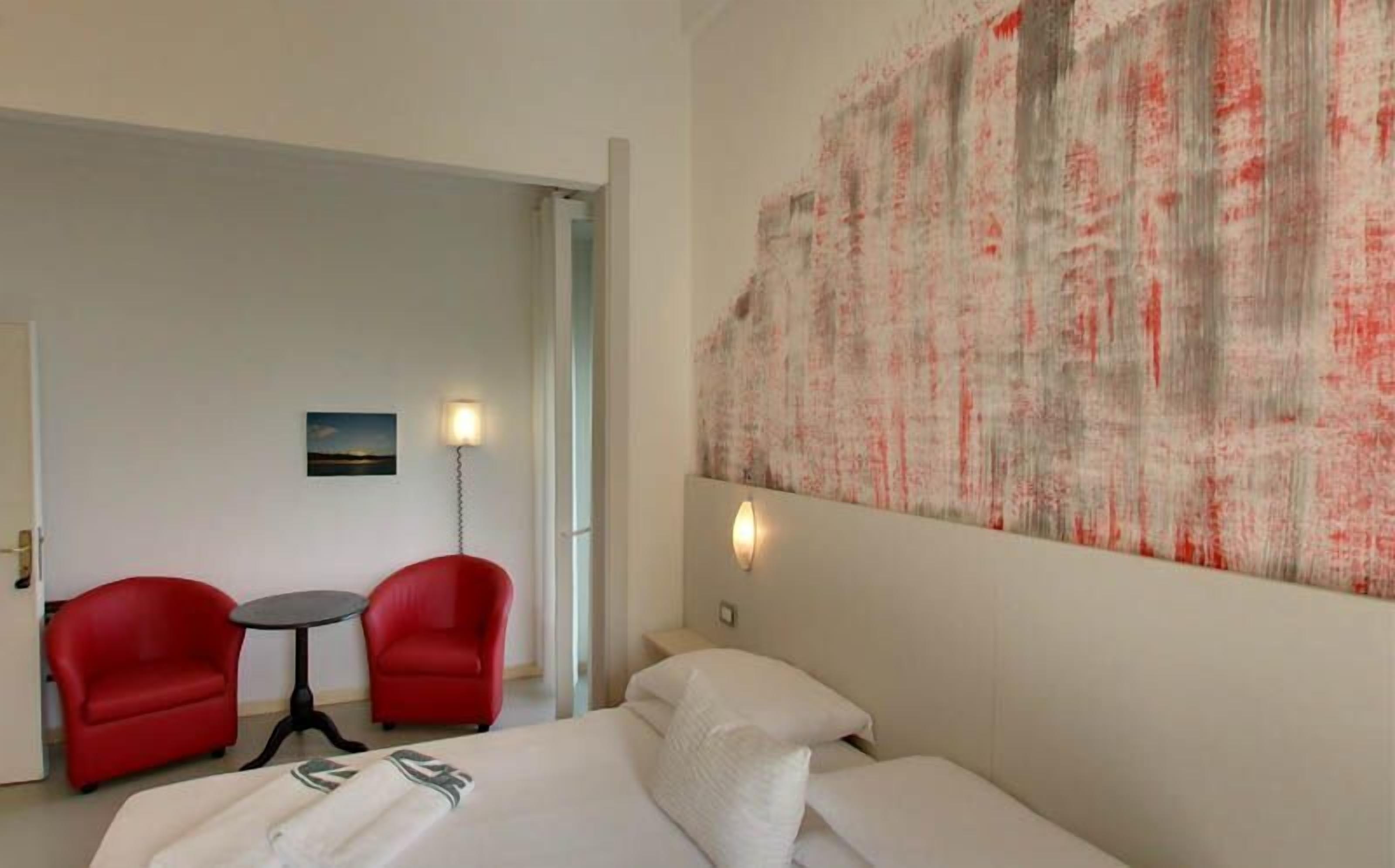 A hotel room with a bed, two red chairs, a small table, and a large abstract painting on the wall.