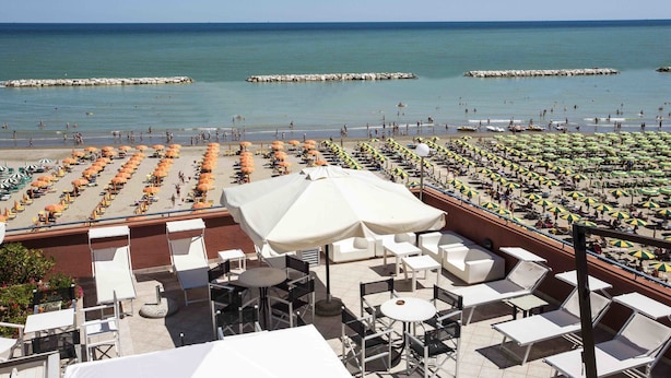 Une terrasse au bord de la plage, dotée de sièges extérieurs et de parasols, qui offre une vue sur une plage animée, remplie de monde et de bateaux.