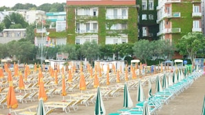 Une plage bordée de rangées de chaises longues et de parasols, une rangée de bâtiments dotés de balcons, et un drapeau en arrière-plan.