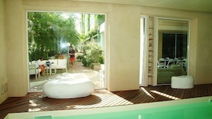 Un espace piscine intérieur moderne, doté d’une terrasse en bois, d’un fauteuil en forme de sac de haricots blanc et d’une porte vitrée donnant sur un espace de détente extérieur.