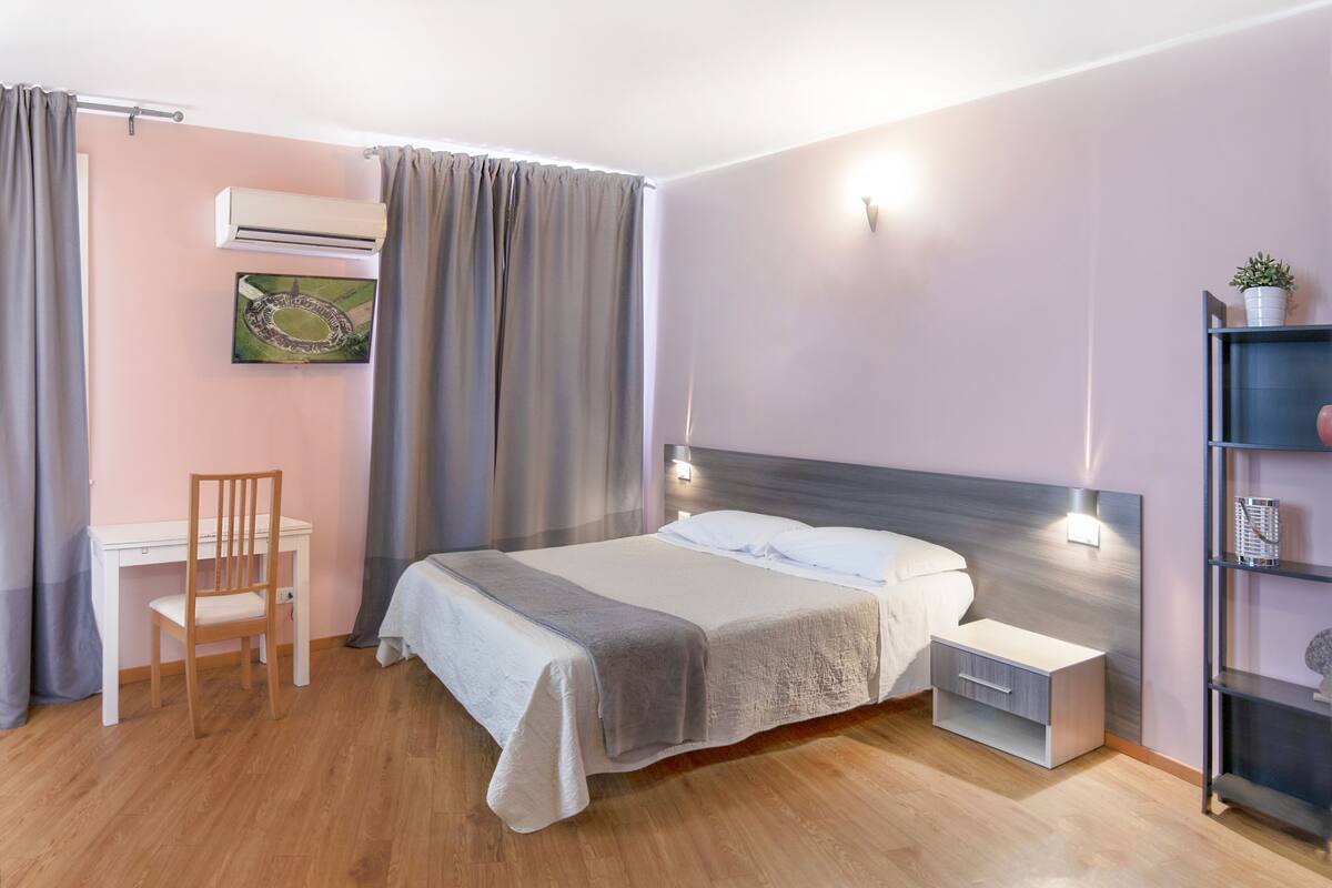 A bedroom with a bed, bedside table, chair, desk, and an air conditioning unit.