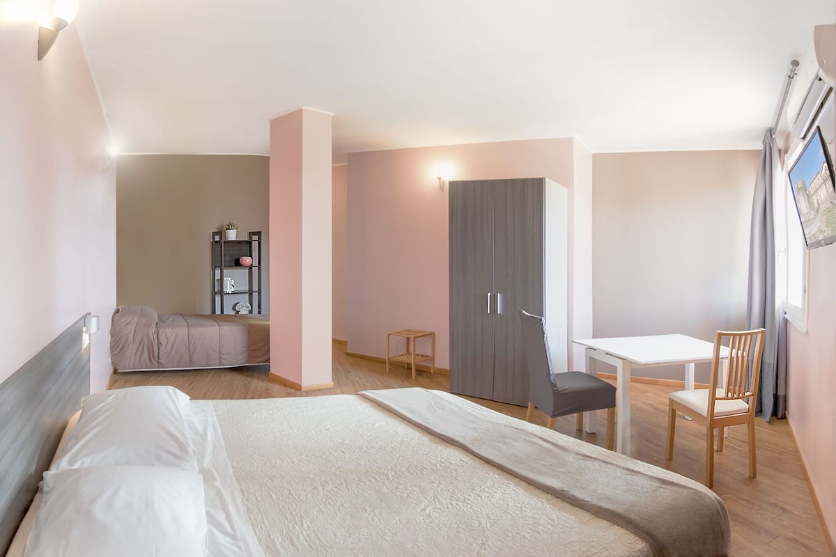 A hotel room with a bed, a desk, a chair, a wardrobe, and a shelf with decorative items.