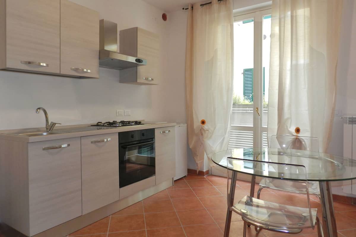 A modern kitchen with a gas stove, oven, and stainless steel cabinets. There is a glass table with chairs and a window with curtains.